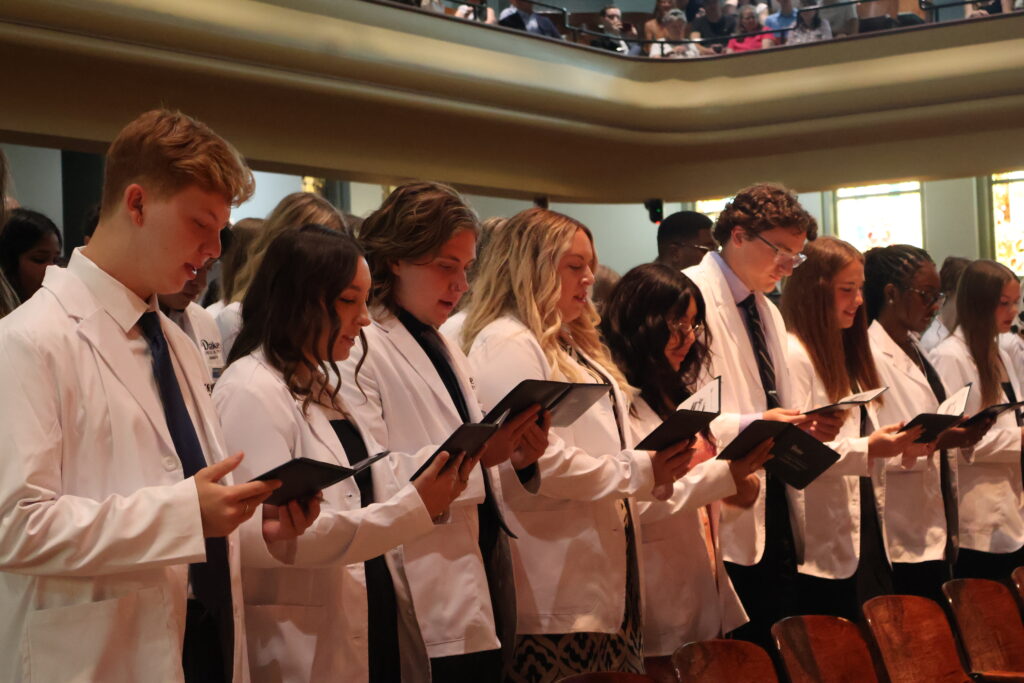 Pharmacy students stand reciting the pharmacy oath together, reading from diplomas in their white coats