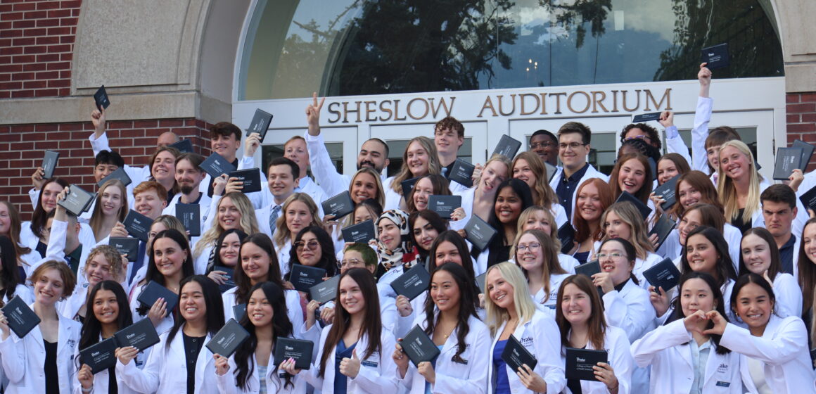 Pharmacy students in group photo wearing white coats and holding up their diplomas