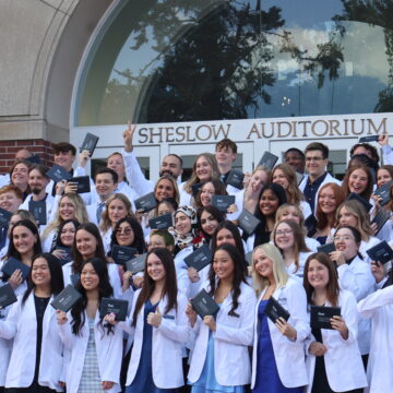 Pharmacy students in group photo wearing white coats and holding up their diplomas