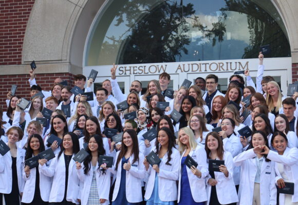 Pharmacy students in group photo wearing white coats and holding up their diplomas