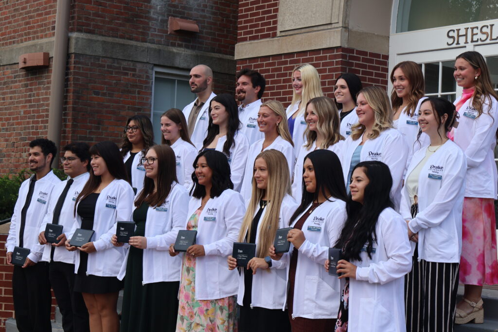 Nursing students stand on stairs and pose for a group photo, holding diplomas and wearing white coats